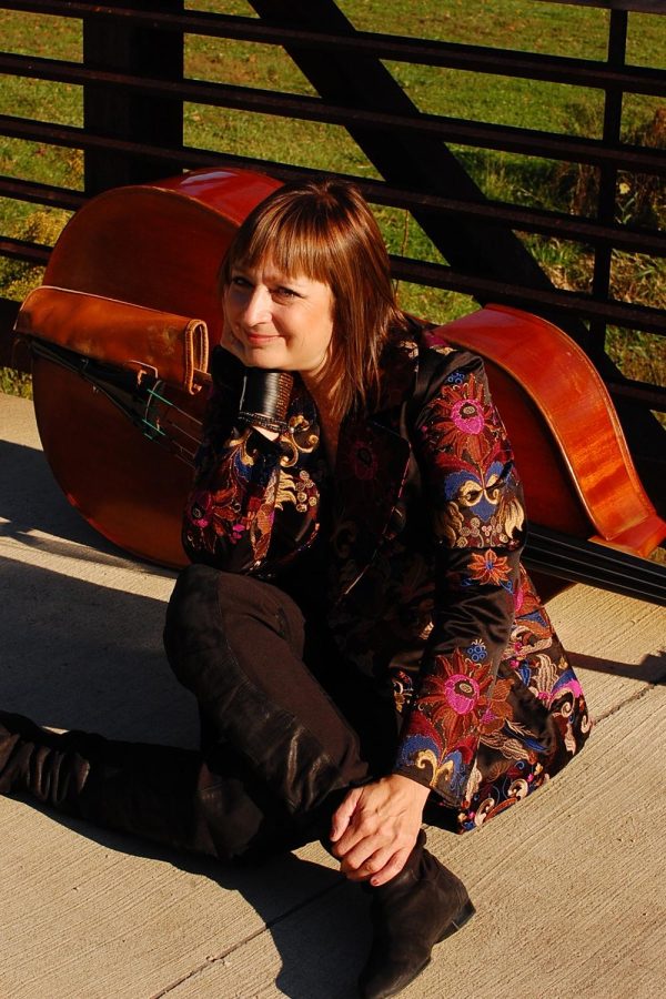 Portrait of a performer sitting with her legs crossed on a sidewalk wearing a floral patterned jacket. A cello is laid on its side behind her; a rustic fence and green grass behind them.