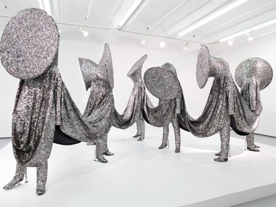Seven human figures with brass instrument heads are entirely covered in a shiny silver sequined fabric that connects each of them.
