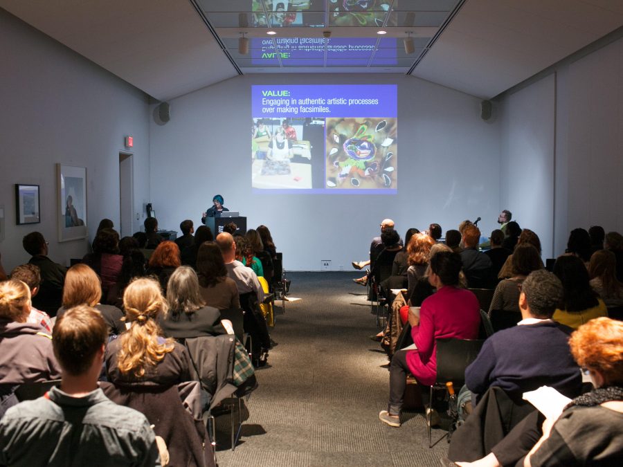 A room is full of people sitting in rows of chairs, taking notes, and looking up at a projection of a presentation, while someone speaks at the front podium.