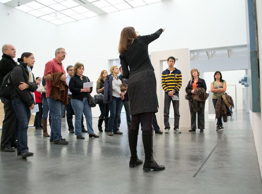 A woman points out a detail in an artwork as a group of people surround her listening and looking at the artwork.