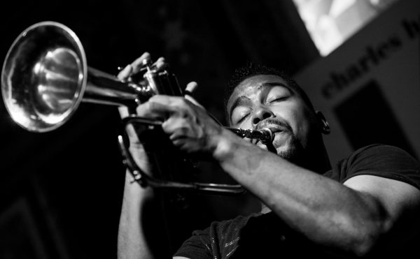 A dark-skinned person with mustache and goatee plays a trumpet with their eyes closed.