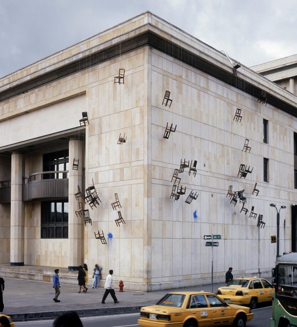 A building has several dozen chairs hanging off its roof and down the side, suspended by rope.