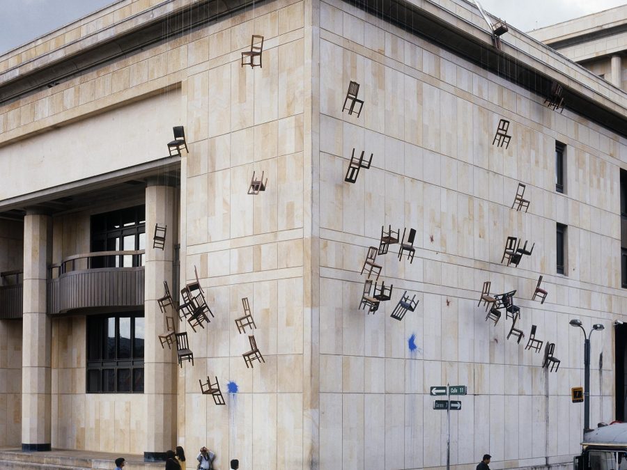 A building has several dozen chairs hanging off its roof and down the side, suspended by rope.