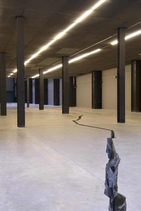 A photograph shows a room with several black columns and a crack in the floor running between them.