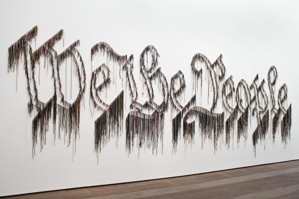 Colorful strands hung on a white wall spell out the phrase 