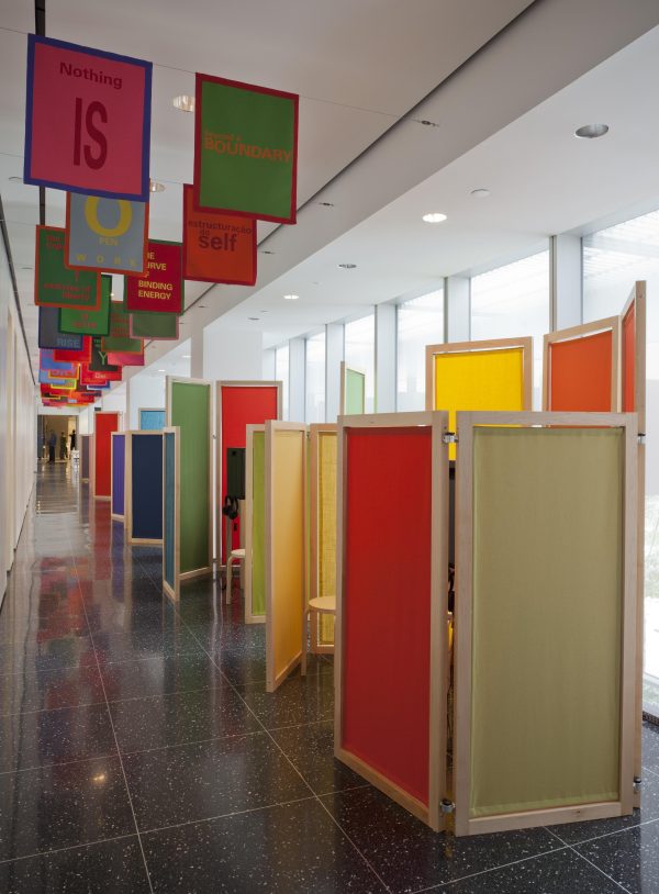 Multicolored banners with text are hung in pairs and wooden and fabric room separators run the length of a white gallery.