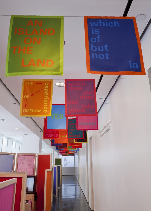 Multicolored banners with text are hung in pairs from the ceiling. Below, wooden and fabric room separators run the length of a white gallery.