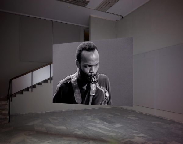 A portrait of a man playing saxophone is seen on a screen suspended from wires in the middle of a room.