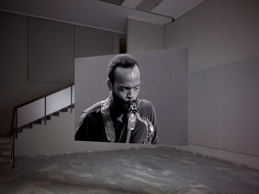A portrait of a man playing saxophone is seen on a screen suspended from wires in the middle of a room.
