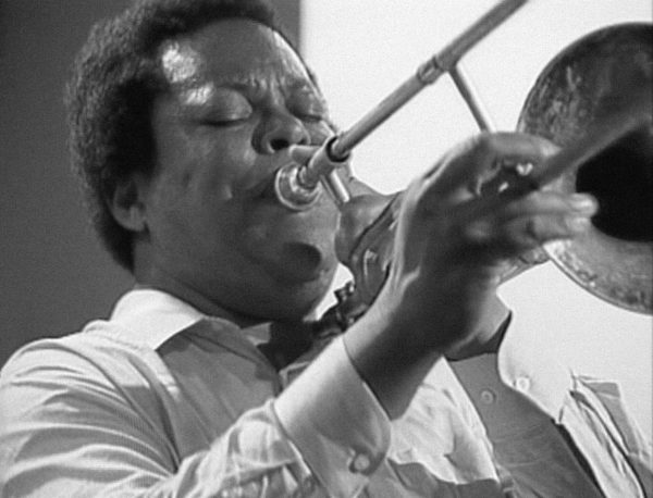 A black-and-white portrait shows a dark-skinned man with a short afro playing the trombone.