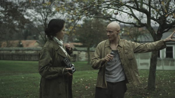 In an outdoor setting, a young bald person reaches their left arm out while looking at a feminine person wearing a trench coat and holding a camera.
