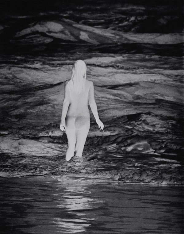 A photo negative of a person with long hair wading knee deep in water that shimmers with light