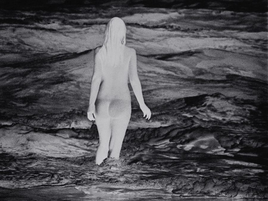 A photo negative of a person with long hair wading knee deep in water that shimmers with light