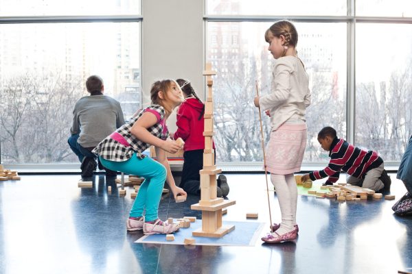 In an open indoor space, children play with wooden building blocks on the floor.