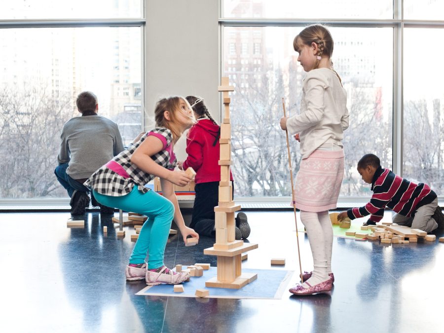 In an open indoor space, children play with wooden building blocks on the floor.