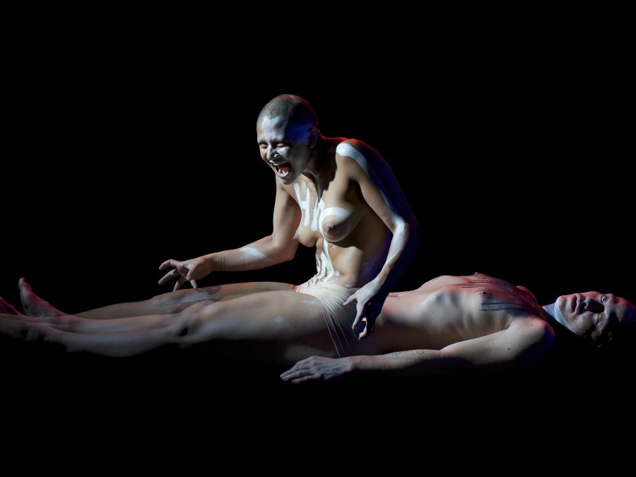 A topless female painted in white lines on their body rises from the base of a shirtless male lying on the ground in a dark space, where only their bodies are lit. The female has a sinister look on their face.