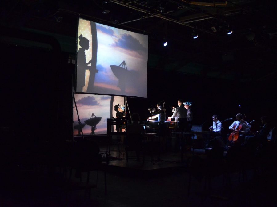 On a dark stage, technicians use overhead projectors to cast ta woman's silhouette onto an image of a sunset that appears in the background.