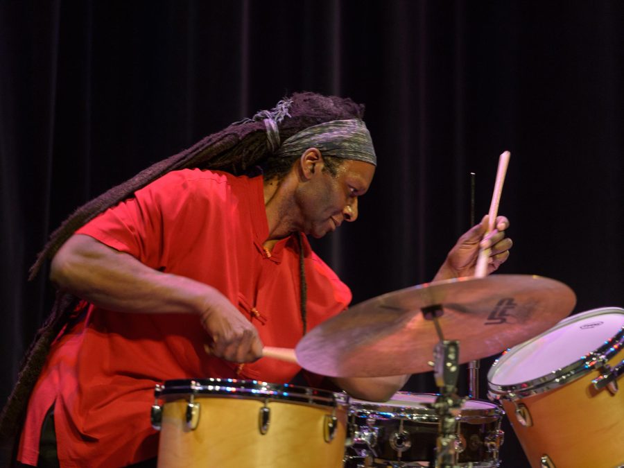 A man with dreadlocks that hang past his waist playing a drum kit in front of a black curtain