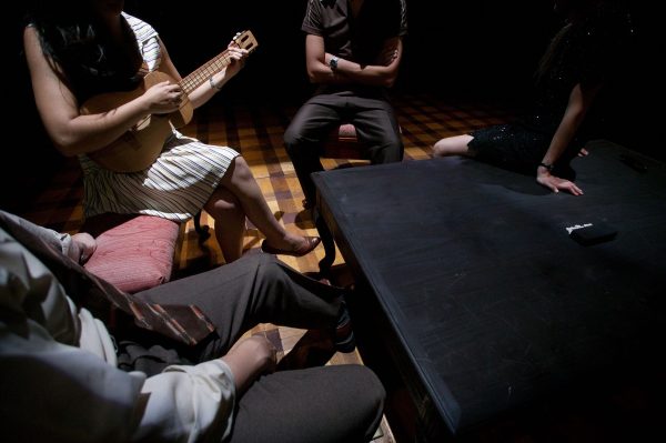 A closely cropped image of four people sitting around a table, one of them is playing a small guitar, we can't see their faces