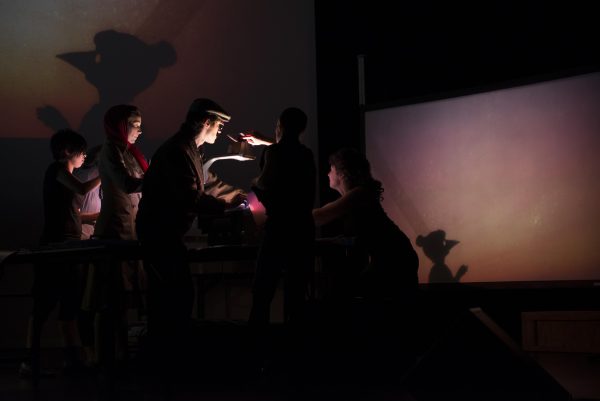 Four technicians use overhead projectors to cast the silhouette of a puppet onto a large screen in the background.