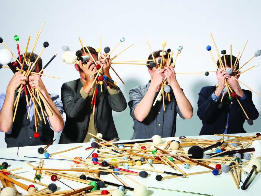 Four men in dress shirts and suit jackets hold handfuls of drumsticks and mallets in front of their faces while sitting at a table also covered with drumsticks and mallets.