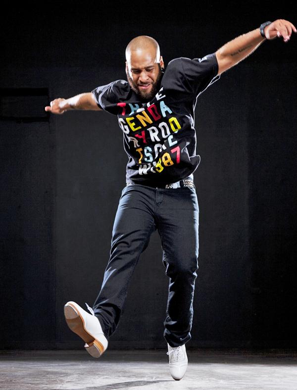 A bald person wearing a mostly black outfit enthusiastically jumps against a black background.