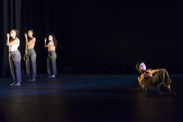 On a dark stage, three women stand in a retreating row on the left and a fourth reclines on the right
