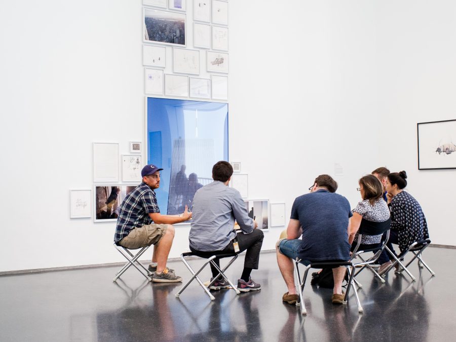 Six people sit on folding stools in front of an installation of drawings and photographs in an arched white room.