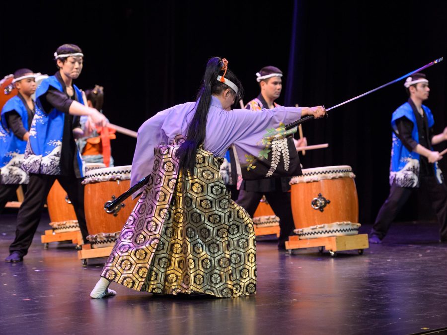 On stage, a woman in traditional Japanese clothing lunges with a long-handled sword while young male drummers play *taiko* drums behind her.