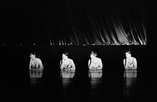 Black-and-white performance still of a dark stage with four dancers visible from the waist up, each resting her chin on a hand