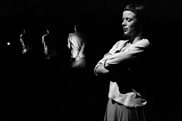 Black-and-white performance still of four dancers progressively disappearing into their pitch-black surroundings