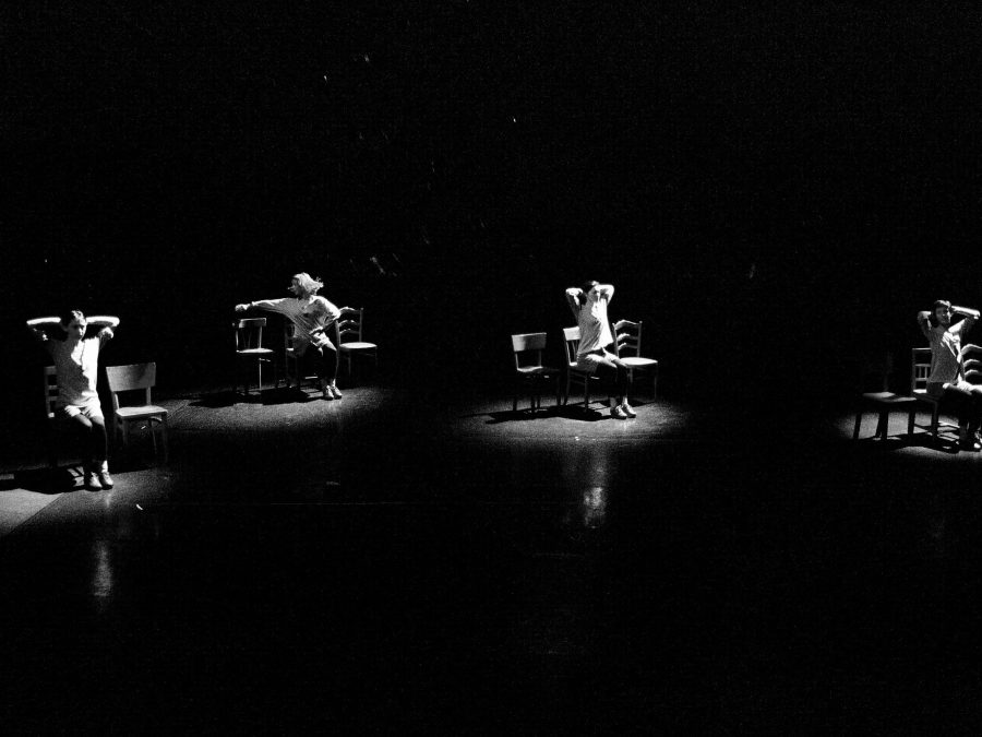 Black-and-white performance still of four women on a pitch-black stage, each one illuminated by a spotlight and dancing in a chair