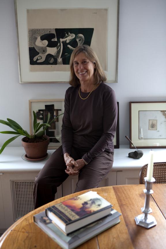 A smiling woman in a brown long-sleeve shirt and pants leans against a white bureau, behind a table stacked with books.