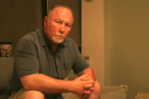 A muscular man with receding hairline and a gray goatee sits, hands folded, looking at the camera, expressionless.
