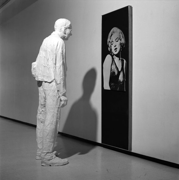 A black-and-white photo shows a white plaster figure of a man in work clothes stands facing a large, vertical, rectangular photograph of an extremely attractive blond woman.