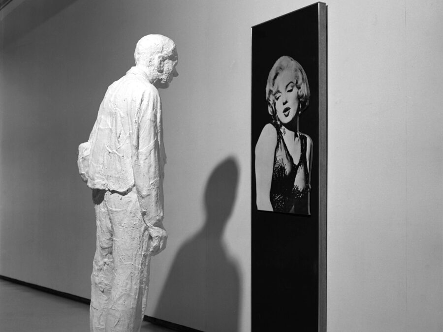 A black-and-white photo shows a white plaster figure of a man in work clothes stands facing a large, vertical, rectangular photograph of an extremely attractive blond woman.