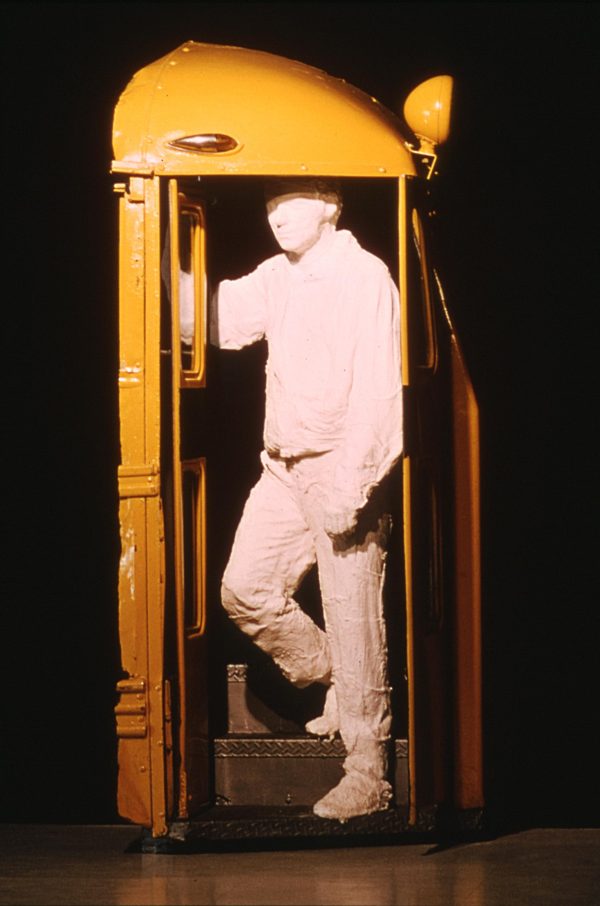 A life-size, white plaster cast of a man in work clothes appears to descend the front step of a yellow school bus.