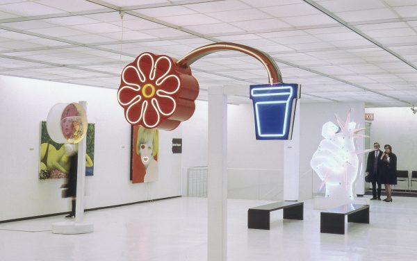 Colorful pop-art sculptures dispersed throughout a white gallery with a low ceiling. The sculpture closest to the foreground is a red flower bending out of a small blue pot.