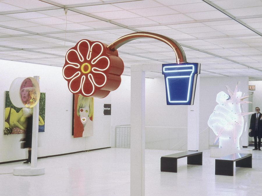 Colorful pop-art sculptures dispersed throughout a white gallery with a low ceiling. The sculpture closest to the foreground is a red flower bending out of a small blue pot.