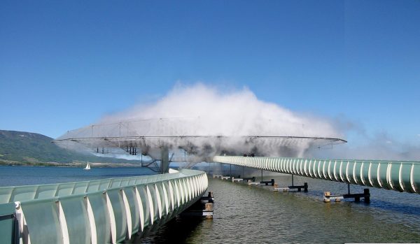 Two floating bridges lead to a saucer-shaped building, engulfed by mist, in the middle of a lake.