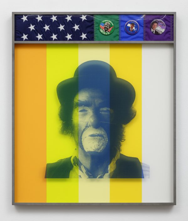 The black-and-white photo of a clown is covered by broad transparent yellow stripes and framed. The top section of the frame displays a blue flag with white stars and three pins featuring different US politicians.