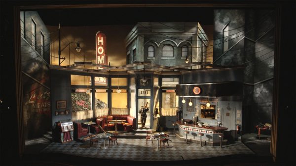 A shadowy stage set featuring a 1950s diner amid row houses, with a single spotlit performer