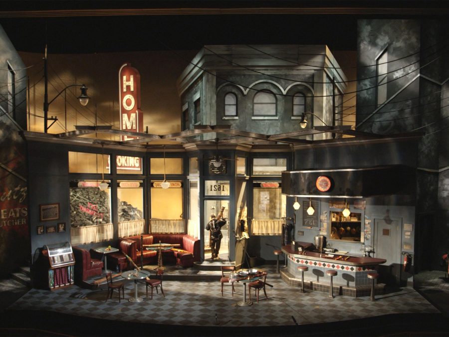 A shadowy stage set featuring a 1950s diner amid row houses, with a single spotlit performer