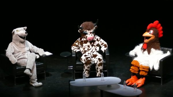 Three people dressed in animal costumes—a rat, a cow, and a chicken—sit on a stage.