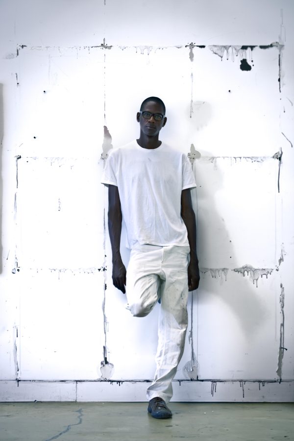 A man with dark brown skin dressed in white posing with one leg up against a white wall featuring a messy gridded outline