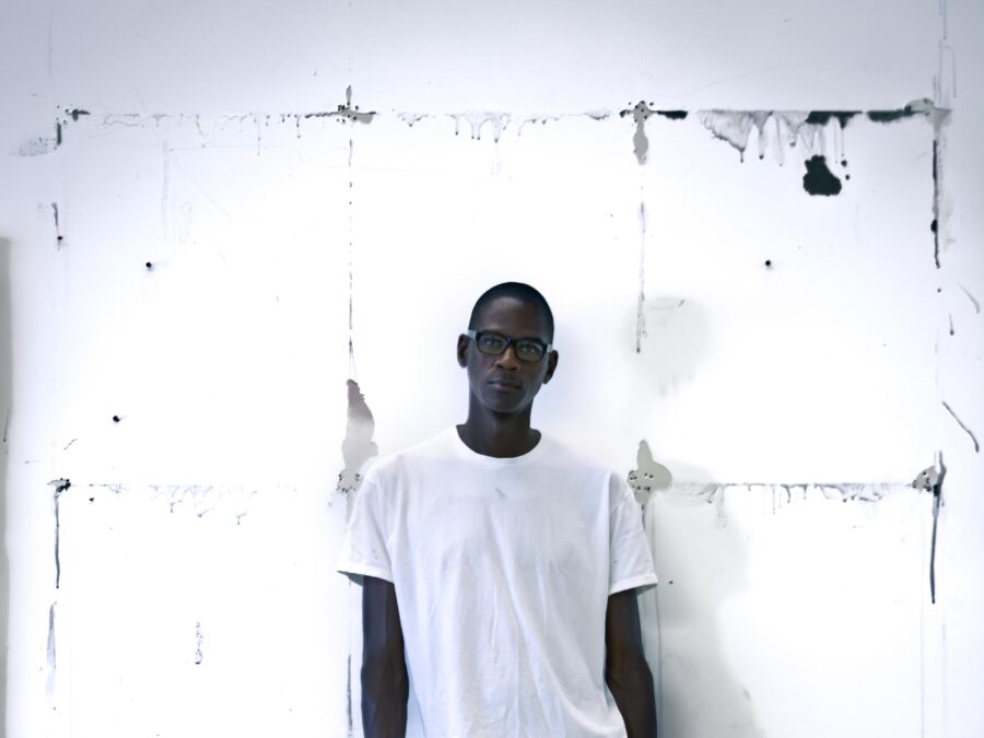 A man with dark brown skin dressed in white posing with one leg up against a white wall featuring a messy gridded outline