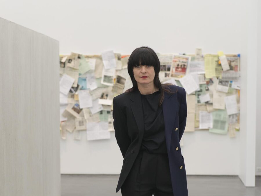 Portrait of a woman with long brown hair, blunt bangs, red lipstick and dressed in black standing in an exhibition at the MCA.