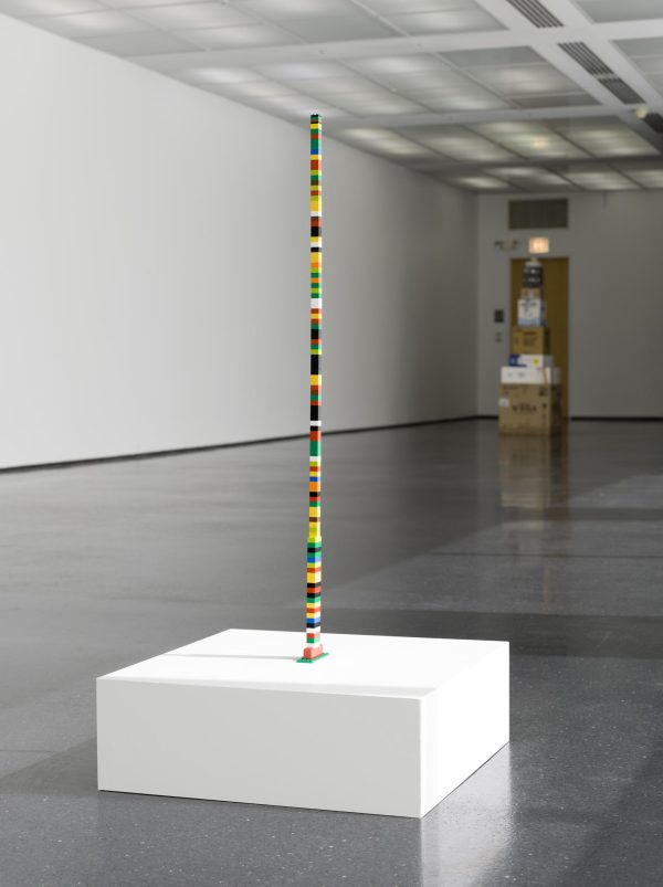 A tower of square building blocks of various colors on a white pedestal in a gallery space