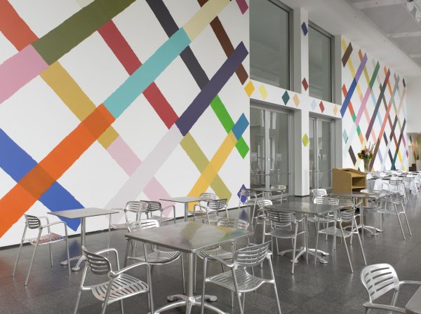 Painted lines of all different colors in a lattice pattern on a white wall in a two-story room with cafe tables