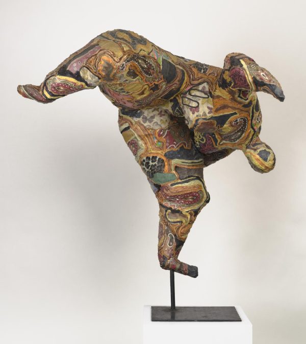 A colorfully patterned figure with large legs and a small head positioned on a metal stand, secured by one leg. The other leg is raised up and out as its head tips back, seemingly doing a somersault in midair.